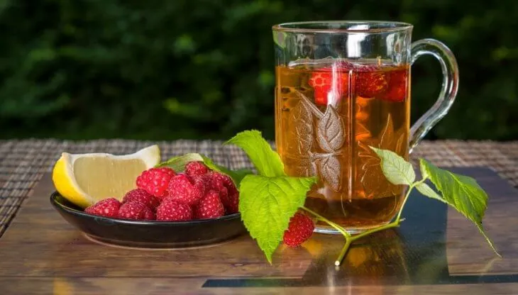 Стеклянная кружка чая, рядом свежая малина и дольки лимона