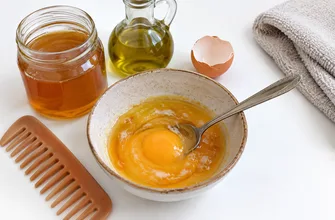 Миска с маской из меда, желтка и оливкового масла, рядом банка меда, масло, яичная скорлупа, гребень и полотенце