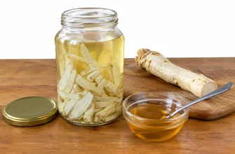 Банка хреновухи с нарезанным корнем хрена, рядом корень хрена на доске и пиала с медом